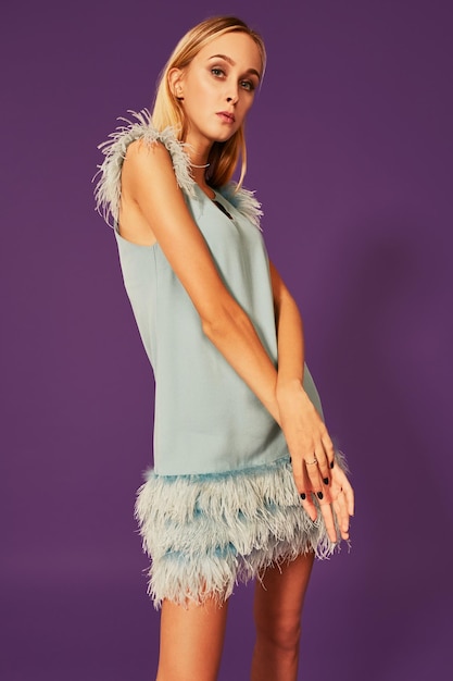 Jeune femme séduisante aux cheveux longs blonds, vêtue d'une élégante robe de cocktail avec des plumes, posant au studio devant la caméra avec les mains croisées