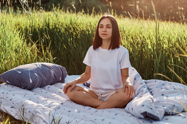 Jeune femme séduisante aux cheveux noirs assise dans une pose de lotus dans un lit moelleux dans un champ vert et méditant, femme portant un t-shirt blanc de style décontracté, regardant à distance exprimant le calme.