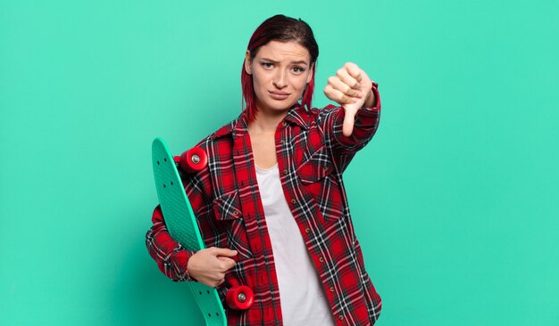 Jeune femme séduisante aux cheveux roux se sentant croisée, en colère, agacée, déçue ou mécontente, montrant les pouces vers le bas avec un regard sérieux et tenant une planche à roulettes