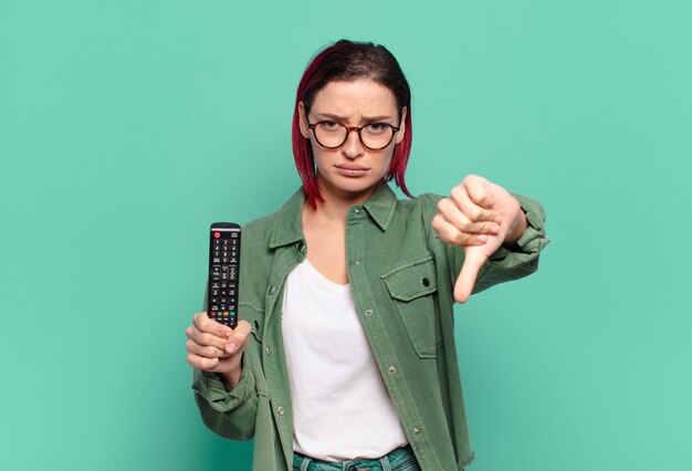 jeune femme séduisante aux cheveux roux se sentant croisée, en colère, agacée, déçue ou mécontente, montrant les pouces vers le bas avec un regard sérieux et tenant une télécommande de télévision