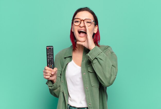 Jeune femme séduisante aux cheveux roux se sentant heureuse, excitée et positive, donnant un grand cri avec les mains à côté de la bouche, appelant et tenant une télécommande de télévision