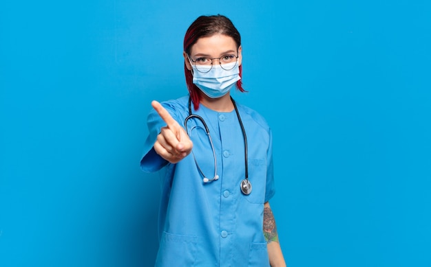 Jeune femme séduisante aux cheveux roux souriant fièrement et en toute confiance faisant la pose numéro un triomphalement, se sentant comme un leader. concept d'infirmière d'hôpital