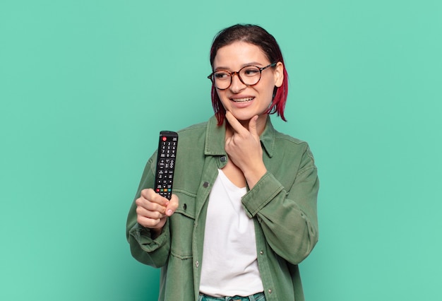 Jeune Femme Séduisante Aux Cheveux Roux Souriante Avec Une Expression Heureuse Et Confiante Avec La Main Sur Le Menton, Se Demandant Et Regardant Sur Le Côté Et Tenant Une Télécommande De Télévision