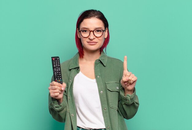 Jeune femme séduisante aux cheveux roux souriante et semblant amicale, montrant le numéro un ou le premier avec la main en avant, comptant à rebours et tenant une télécommande de télévision