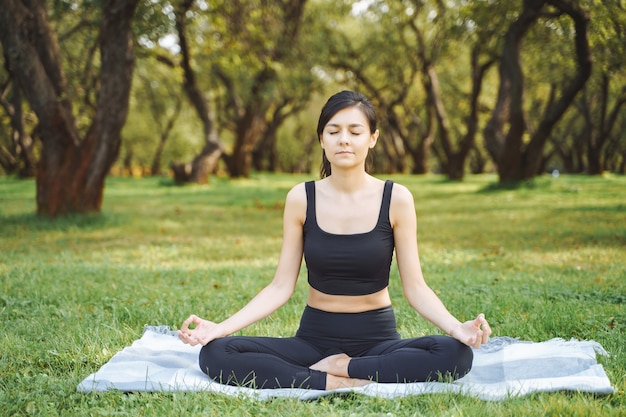 Jeune femme séduisante aux yeux fermés méditant en position du lotus assis sur l'herbe dans le parc.