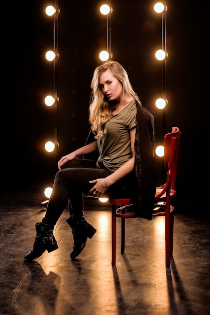 Jeune femme séduisante blonde en t-shirt et costume avec verre assis sur la chaise sur la scène