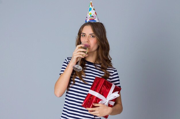 Jeune femme séduisante en casquette de célébration tenant un cadeau rouge et buvant une coupe de champagne