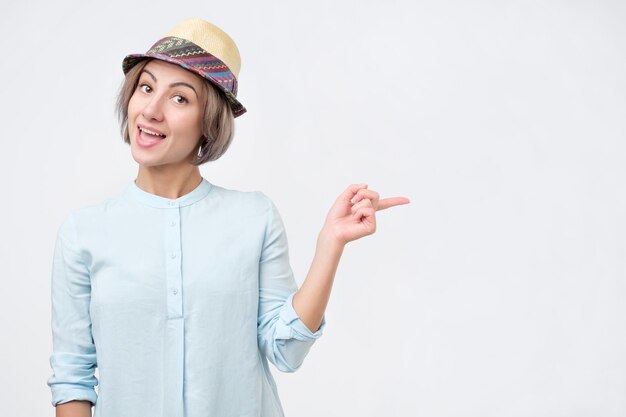 Jeune femme séduisante en chapeau d'été et chemise bleue pointant avec son doigt sur le fond