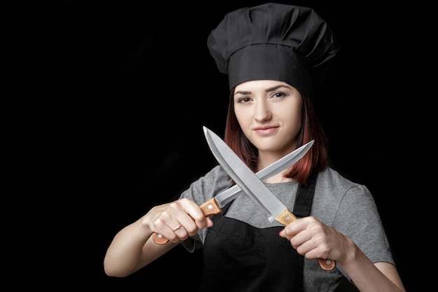 Jeune femme séduisante chef en uniforme noir tient deux couteaux sur fond noir