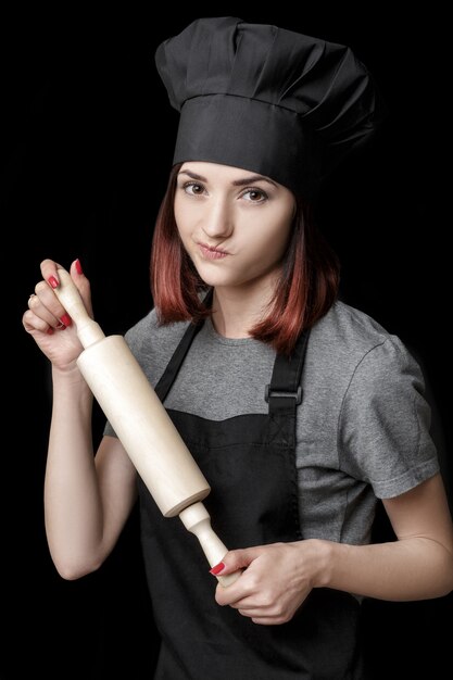 Jeune femme séduisante chef en uniforme noir tient un rouleau à pâtisserie sur fond noir