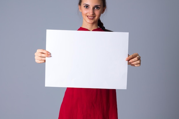 Jeune femme séduisante en chemisier rouge avec tresse montrant une feuille de papier blanche sur fond gris en studio