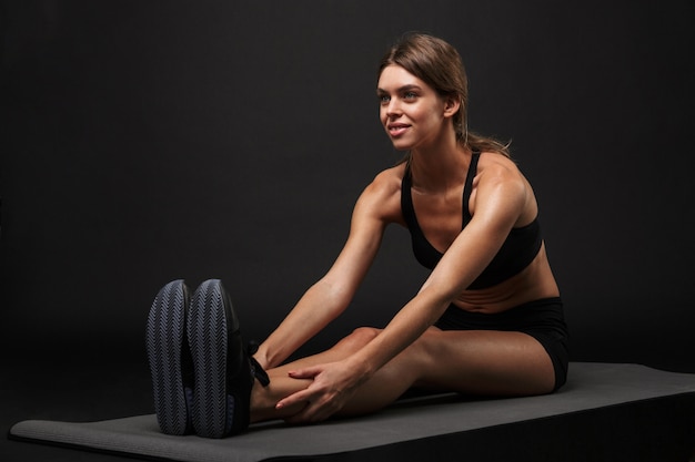 Jeune femme séduisante et confiante en bonne santé portant un soutien-gorge de sport et un short isolé sur fond noir, s'exerçant sur un tapis de fitness, étirement de yoga