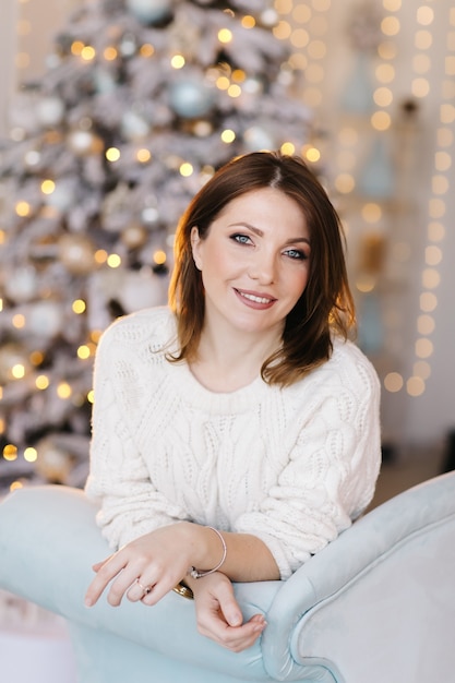 Une jeune femme séduisante dans un pull blanc est assise sur le canapé près de l'arbre de Noël