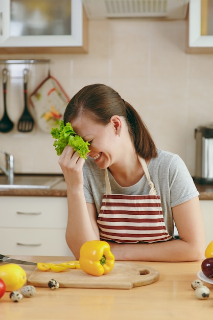 La jeune femme séduisante dans un tablier avec des feuilles de laitue riant dans la cuisine. Concept de régime. Mode de vie sain. Cuisiner à la maison. Préparer la nourriture.