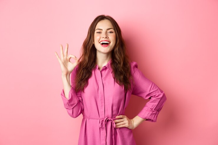 Jeune femme séduisante disant oui, montrant un bon signe d'approbation, souriant confiant et satisfait, debout sur un mur rose.