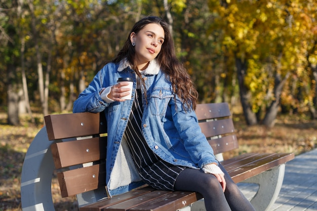 Une jeune femme séduisante est assise sur un banc dans un parc en automne avec une tasse de café dans les mains