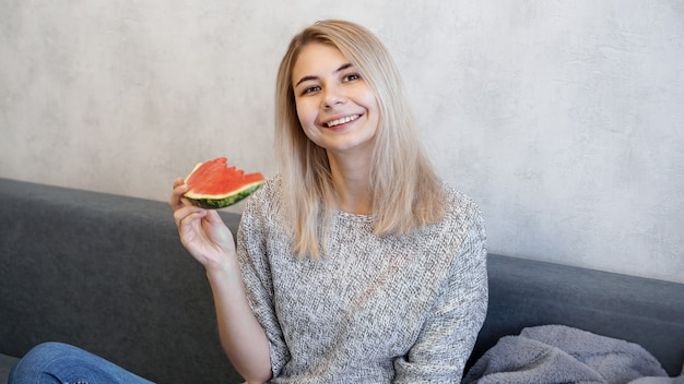 Jeune femme séduisante, manger de la pastèque. Femme à la maison dans un intérieur confortable regardant la caméra et souriant