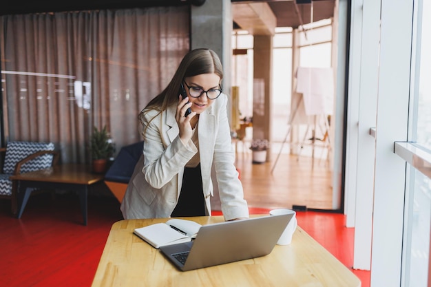 Une jeune femme séduisante parle sur un téléphone portable et sourit tout en étant assise seule près d'une grande fenêtre dans un café pendant son temps libre et en travaillant sur un ordinateur Une femme heureuse se repose dans un café