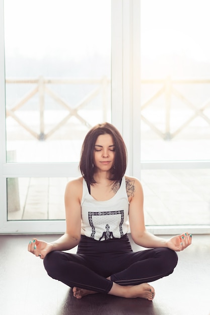 Jeune femme séduisante pratiquant le yoga assis dans une pose de lotus près d'une grande fenêtre avec la lumière du soleil