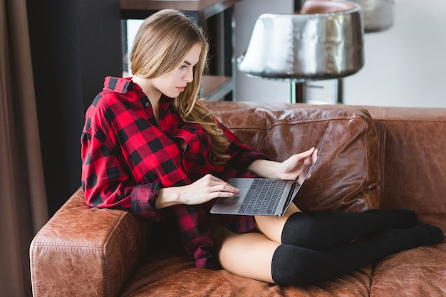 Jeune femme séduisante réfléchie en chemise à carreaux et bas noirs travaillant avec un ordinateur portable sur un canapé en cuir à la maison