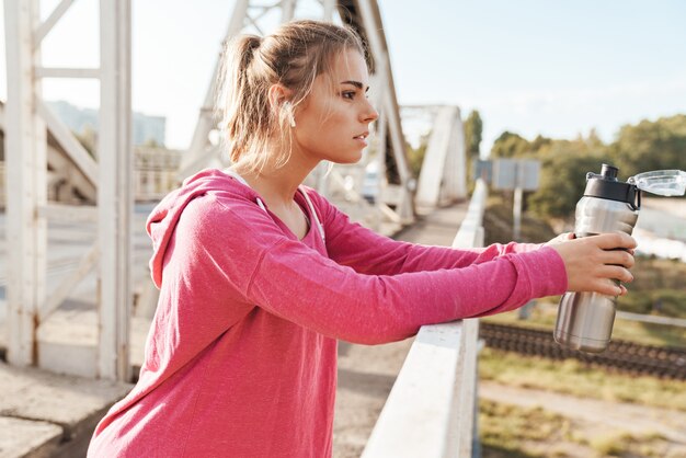 Photo jeune femme séduisante de remise en forme travaillant à l'extérieur sur le pont, écoutant de la musique avec des écouteurs, tenant une bouteille d'eau