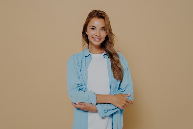 Jeune femme séduisante et satisfaite montrant de la confiance avec les bras croisés et un large et beau sourire sur son visage, l'air positive et inspirée sur fond beige. Expression du visage humain