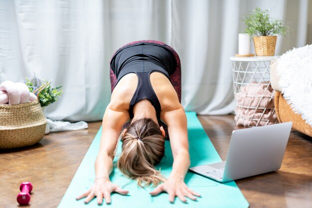 Jeune femme séduisante sportive travaillant à pratiquer le yoga et à l'aide d'un ordinateur portable et d'haltères à la maison en