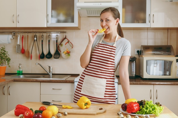 La jeune femme séduisante en tablier goûte le poivre jaune dans la cuisine. Concept de régime. Mode de vie sain. Cuisiner à la maison. Préparer la nourriture.