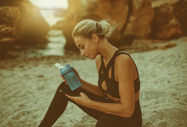 Une jeune femme séduisante en tenue de sport se repose après l'entraînement alors qu'elle est assise sur le sable de la plage sauvage. Une femme sportive utilise un smartphone et boit de l'eau d'une bouteille