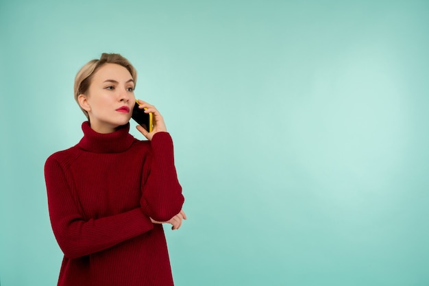 Une jeune femme sérieuse dans un pull rouge avec un smartphone sur fond bleu