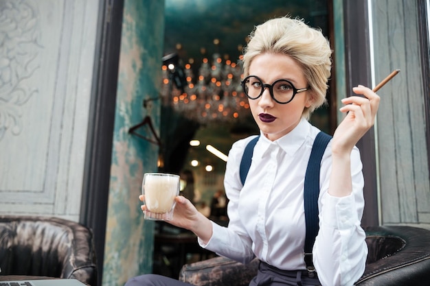 Jeune femme sérieuse dans des verres ronds fumant la cigarette et buvant du café dans le café extérieur