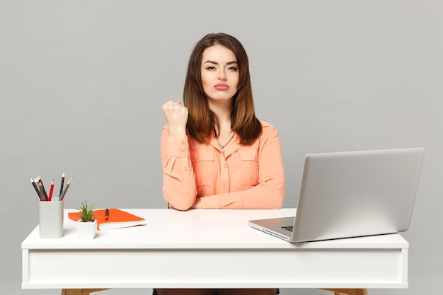 Jeune femme sérieuse dans des vêtements décontractés pastel serrant le poing, s'asseoir et travailler au bureau blanc avec ordinateur portable pc isolé sur fond gris. Concept de style de vie de carrière d'affaires de réalisation. Maquette de l'espace de copie.