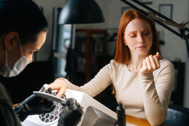 Jeune femme sérieuse examinant les ongles traités et évaluant le travail d'une manucure dans un salon de cosmétologie