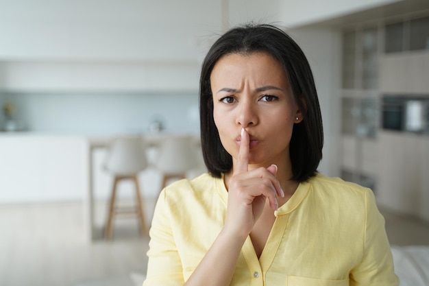 Une jeune femme sérieuse et fronçant les sourcils montre un geste silencieux demandant de garder la vie privée secrète et le silence à la maison