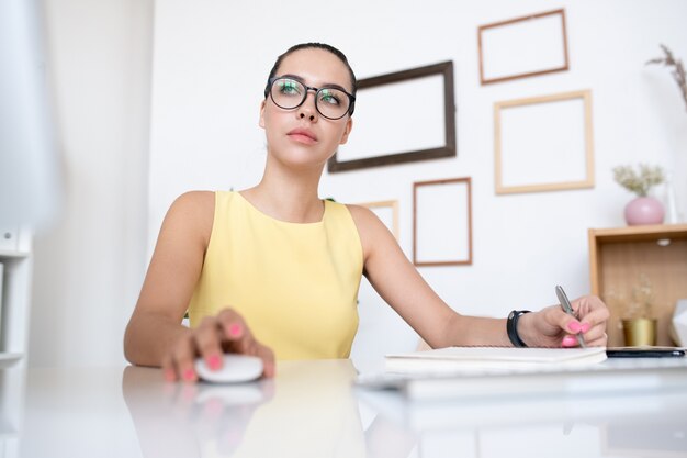 Jeune femme sérieuse gestionnaire de bureau en robe jaune en cliquant sur la souris de l'ordinateur tout en regardant l'écran pendant l'organisation du travail