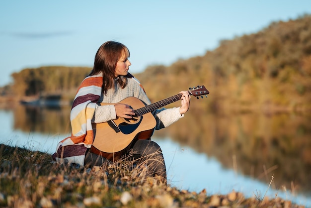 Une jeune femme sérieuse jouant de la guitare sur la rive de la rivière Concept de profiter d'un soir ensoleillé dans la nature heure d'or temps frais et musique
