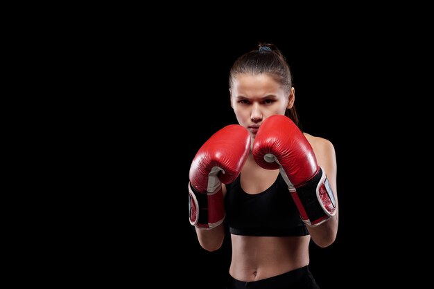 Jeune femme sérieuse et puissante en vêtements de sport et gants de boxe vous regarde prêt à se battre sur fond noir