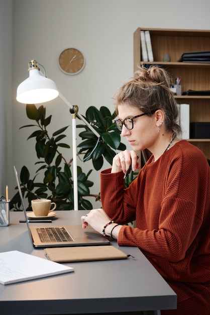 Jeune femme sérieuse se concentrant sur son travail en ligne alors qu'elle était assise à la table et travaillait sur un ordinateur portable