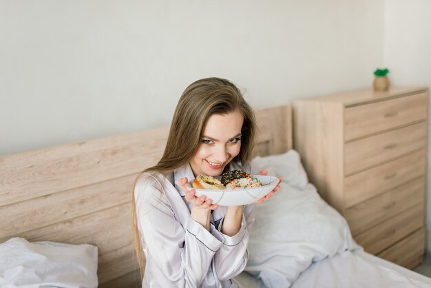 Jeune femme en serviette blanche et robe dans la cuisine pendant la quarantaine. Tenez le beignet sur une assiette. L'heure du petit déjeuner à la maison.