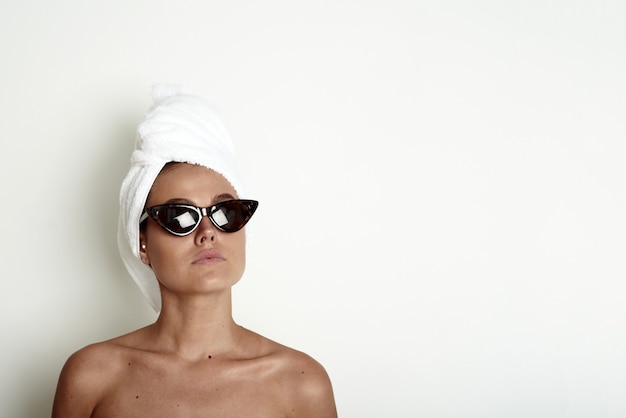 Jeune femme avec une serviette blanche sur la tête et des lunettes de soleil. Concept de soins de la peau du matin, modèle drôle. Copiez l'espace.