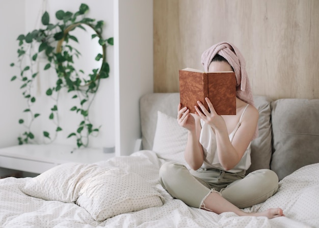 Jeune femme avec une serviette sur la tête lisant un livre au lit le matin