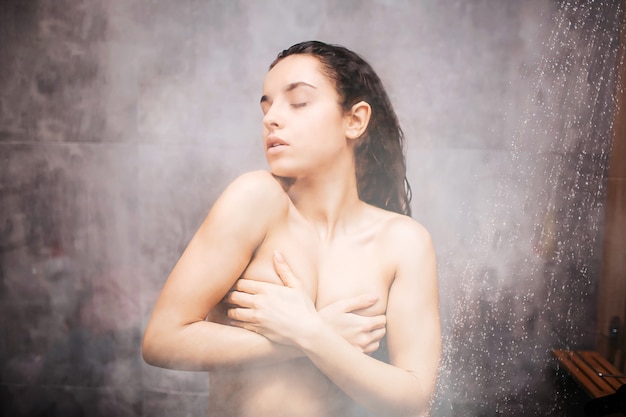 Jeune femme sexy séduisante sous la douche. Plaisir en se lavant. Couvrir la poitrine avec les mains. Yeux fermés. Vapeur d'eau sur mur de verre. Image semi floue.