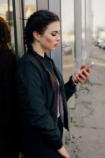 Jeune femme sexy avec des tresses vêtue d'un pantalon noir et d'un blouson noir tenant un téléphone.