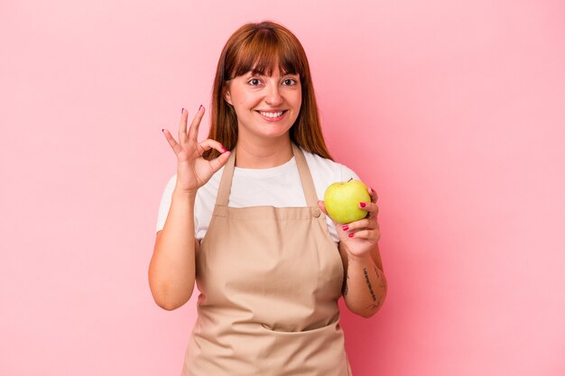 Jeune femme sinueuse caucasienne cuisinant à la maison tenant une pomme isolée sur fond rose joyeuse et confiante montrant un geste ok.