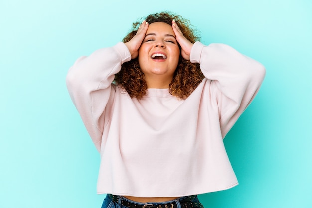 Jeune femme sinueuse latine isolée sur le mur bleu rit joyeusement en gardant les mains sur la tête. Concept de bonheur.