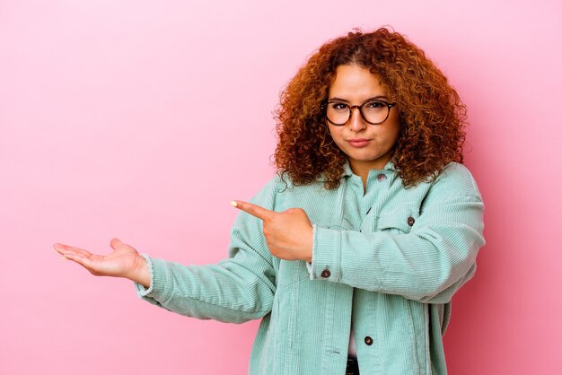 Jeune femme sinueuse latine isolée sur un mur rose excité tenant un espace de copie sur la paume.