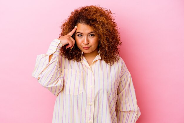 Jeune femme sinueuse latine isolée sur le mur rose pointant le temple avec le doigt, pensant, concentré sur une tâche.