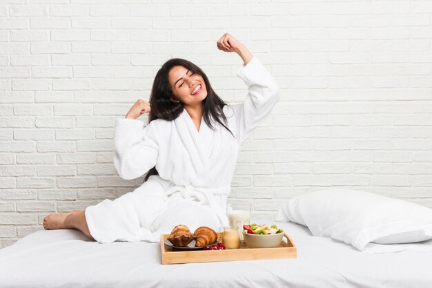 Jeune femme sinueuse prenant un petit déjeuner sur le lit en levant le poing après une victoire, concept gagnant.