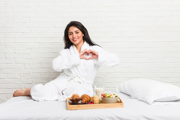 Jeune femme sinueuse prenant un petit déjeuner sur le lit en souriant et montrant une forme de coeur avec les mains.