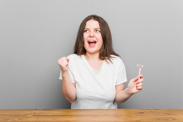 Jeune femme sinueuse de taille plus tenant une lame de rasoir applaudissant sans soucis et excité. Concept de victoire.